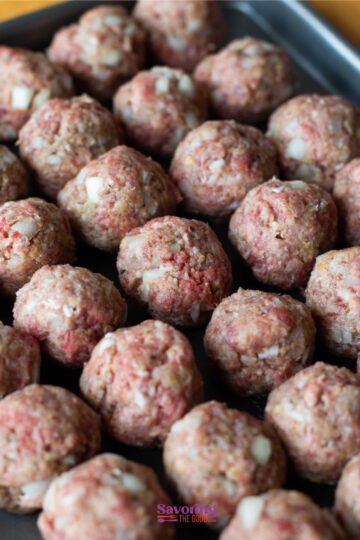 A baking tray filled with rows of uncooked meatballs mixed with chopped onions.