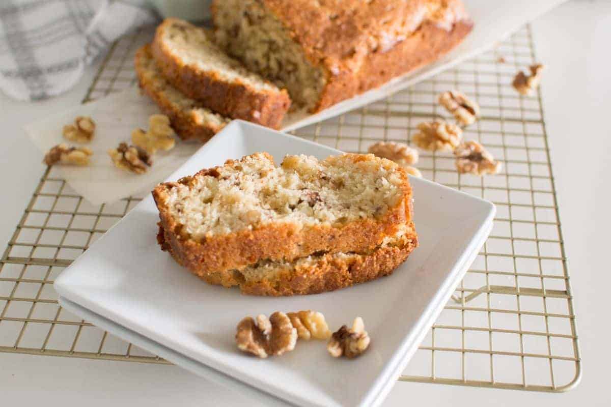 A slice of banana bread on a white plate, garnished with walnut halves, with more sliced loaf cake and walnuts in the background.