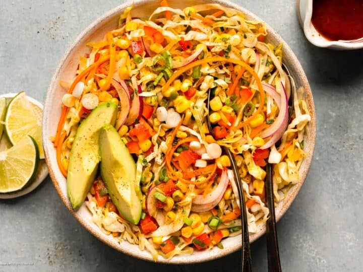 A vibrant salad with sliced avocado, corn, shredded vegetables, red onions, and green onions in a bowl with lime wedges on the side and a small cup of dressing.