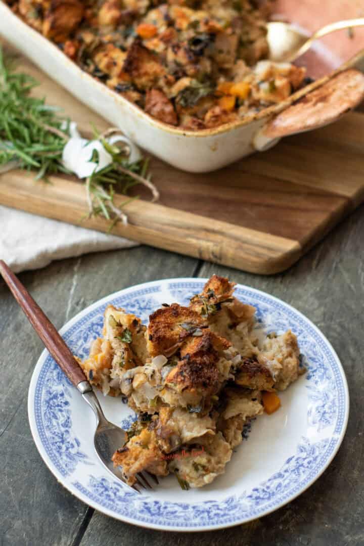 The image shows a plate of stuffing with a fork, garnished with herbs, in front of a dish of more stuffing on a wooden board.