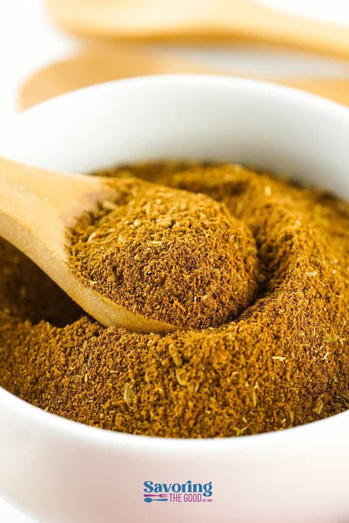 A wooden spoon resting in a white bowl filled with ground spices. The bowl is placed on a white surface.