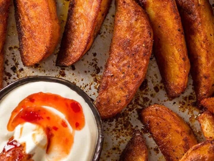 A tray of crispy potato wedges with one wedge dipped in a small bowl of creamy white sauce topped with red sauce. The background shows more wedges on a slightly greasy baking sheet.