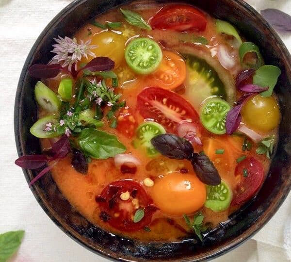 A colorful bowl of gazpacho topped with sliced green and red tomatoes, purple basil leaves, and edible flowers is set against a white background with scattered cherry tomatoes and greens.