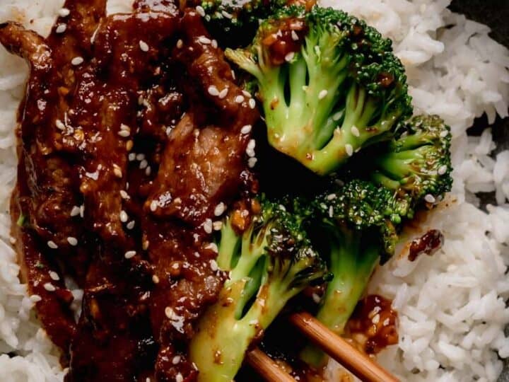 A bowl of white rice topped with beef strips in a dark sauce, broccoli florets, sesame seeds, and chopsticks.