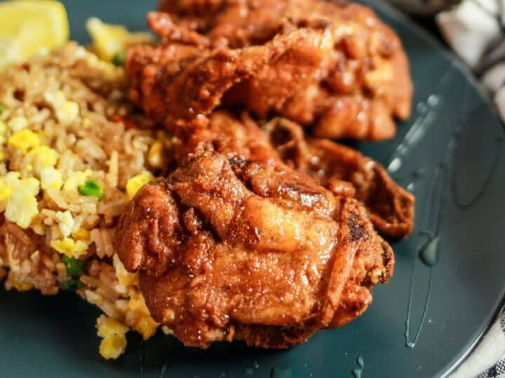 A plate of fried rice with mixed vegetables served alongside two pieces of fried chicken, garnished with a lemon wedge on a dark round plate. A white and black checkered cloth is partially visible.