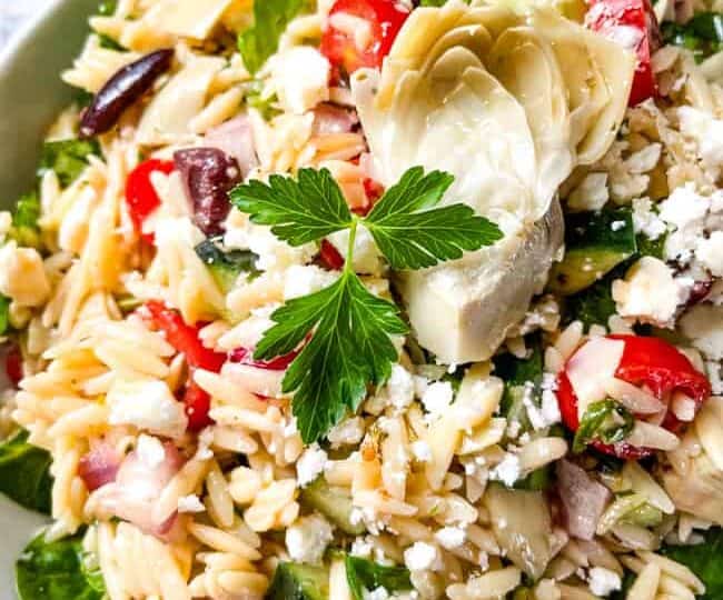 Close-up of a mediterranean orzo pasta salad with cucumbers, tomatoes, olives, red onions, feta cheese, spinach, and an artichoke heart, garnished with a sprig of parsley.