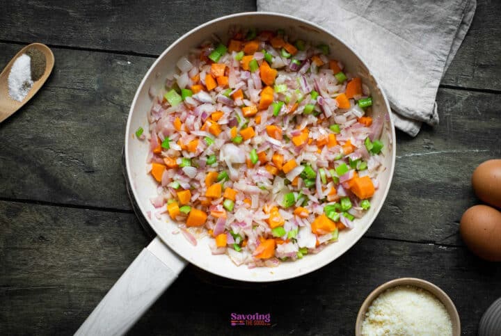A skillet filled with diced onions, carrots, and celery sits on a wooden surface, accompanied by a bowl of grated cheese, two eggs, and a wooden spoon with salt-perfect for crafting your favorite vegetable stuffing recipe.