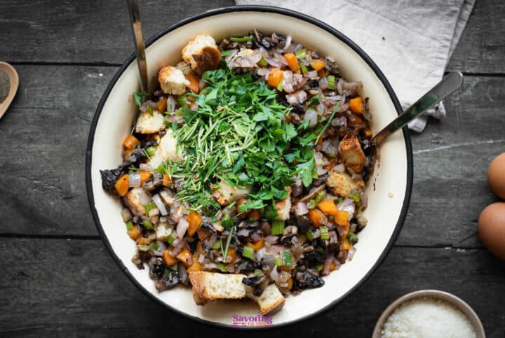 A bowl filled with mixed vegetables, croutons, and fresh herbs, with two spoons in it, rests on a wooden surface. Nearby are two eggs and a small bowl of grated white cheese-perfect ingredients for your next vegetable stuffing recipe.