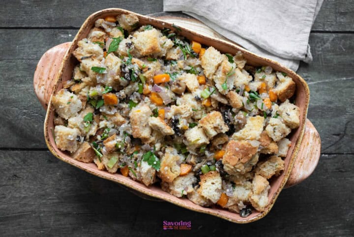 A ceramic baking dish filled with vegetable stuffing recipe featuring an array of vegetables and herbs, placed on a wooden surface. A cloth napkin is partially visible in the background.