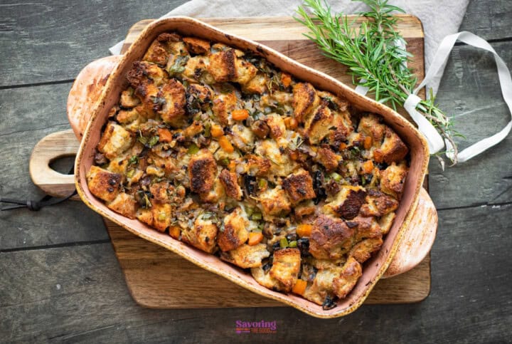 A casserole dish filled with baked bread stuffing, garnished with herbs and chunks of vegetables, displayed on a wooden cutting board. Perfect for those seeking a hearty vegetable stuffing recipe!