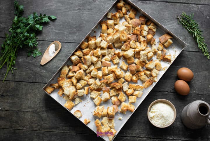 A tray of croutons is positioned in the center on a wooden table, surrounded by fresh herbs, a wooden spoon of salt, two eggs, grated cheese in a bowl, and an oil jar-perfect for your next vegetable stuffing recipe.