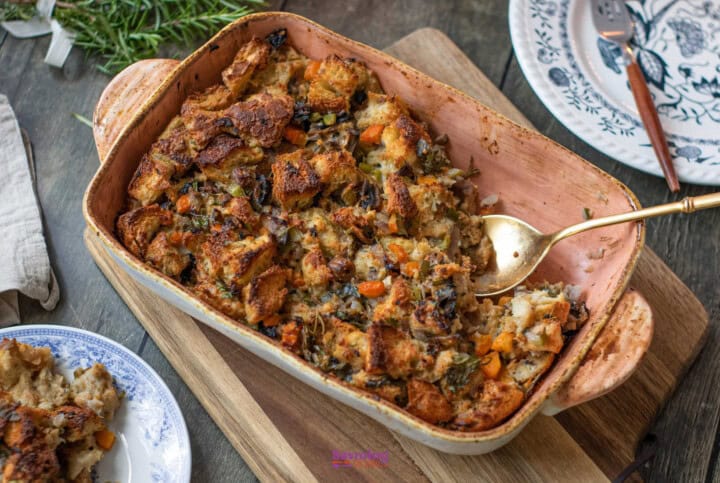 A casserole dish filled with baked bread stuffing containing vegetables such as carrots and herbs, with a serving spoon on the right and a portion served on a blue-rimmed plate in the background showcases this delightful vegetable stuffing recipe.