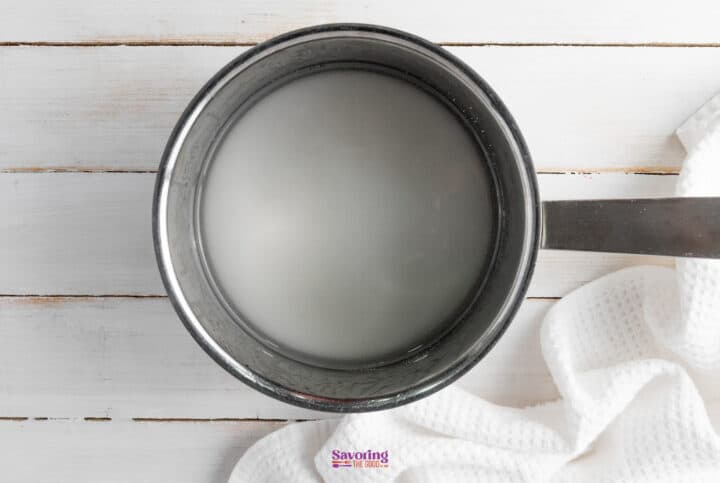 A metal saucepan filled with a white liquid sits on a white wooden surface next to a textured white cloth, hinting at the sweet aroma of strawberry lemonade.