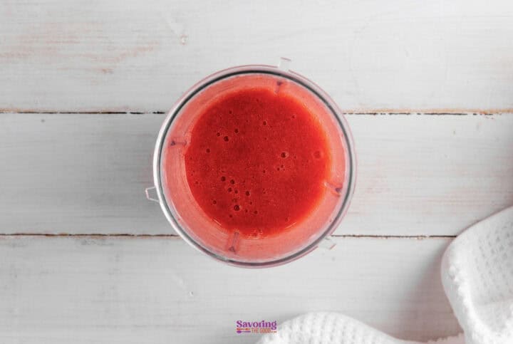 A top view of a blender container filled with a smooth red liquid, likely a strawberry lemonade smoothie, placed on a light wooden surface next to a white cloth.