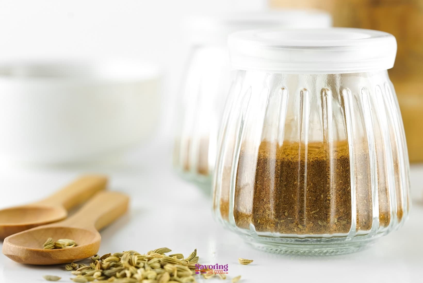 Three glass jars with white lids containing ground spices, placed on a white surface next to wooden spoons and scattered fennel seeds, evoke the rich aroma of Chinese five spice.