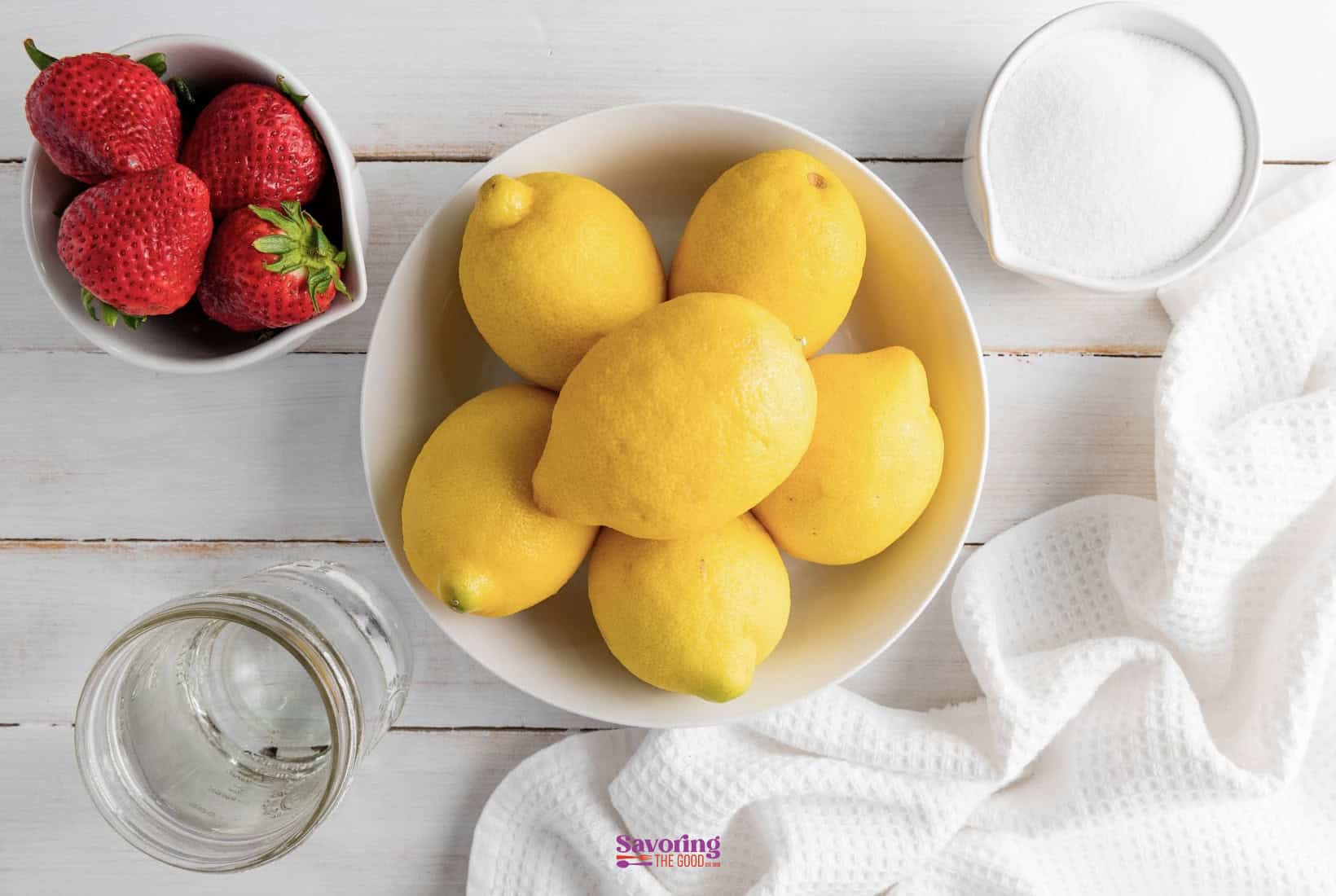 A bowl of lemons, a bowl with strawberries, a glass of water, and a bowl of white sugar are arranged on a white wooden surface with a white cloth nearby.