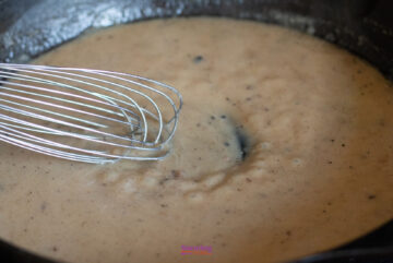 A metal whisk stirs a pan of light brown gravy with visible specks of seasoning, reminiscent of the savory goodness in an IKEA Swedish meatballs recipe.