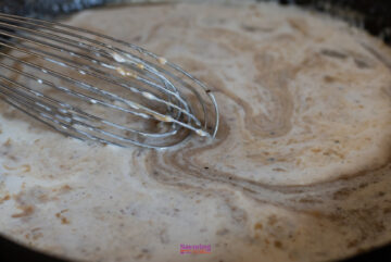 A close-up of a whisk stirring a pot of cream sauce, creating a swirled pattern, reminiscent of preparing an IKEA Swedish meatballs recipe.