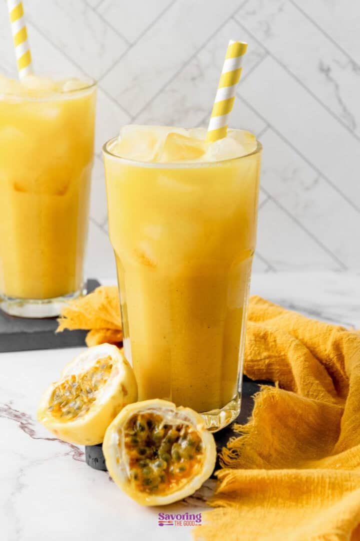 Two tall glasses of passion fruit juice with ice and yellow-striped straws, placed on a marble surface. A halved passion fruit and a yellow cloth are beside the front glass.