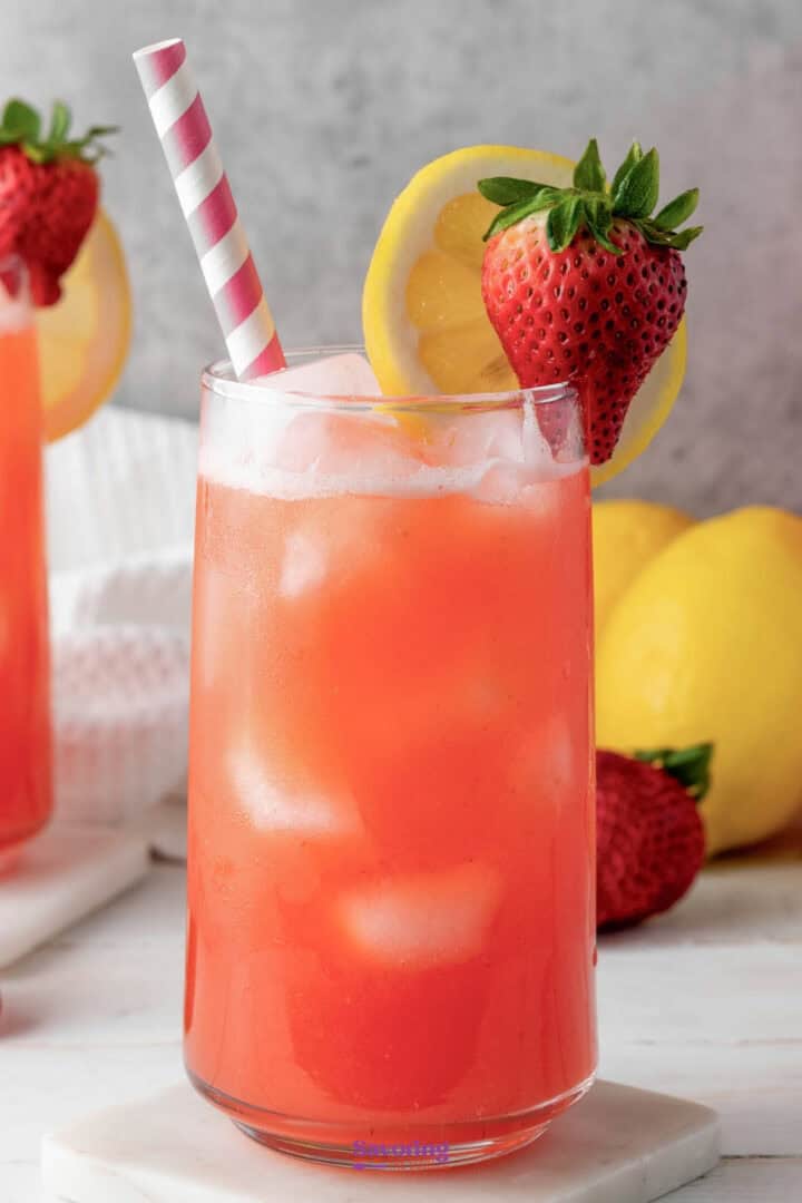 A glass of iced strawberry lemonade garnished with a lemon slice and a strawberry, with a pink and white striped straw. Other lemons and a second glass are visible in the background.
