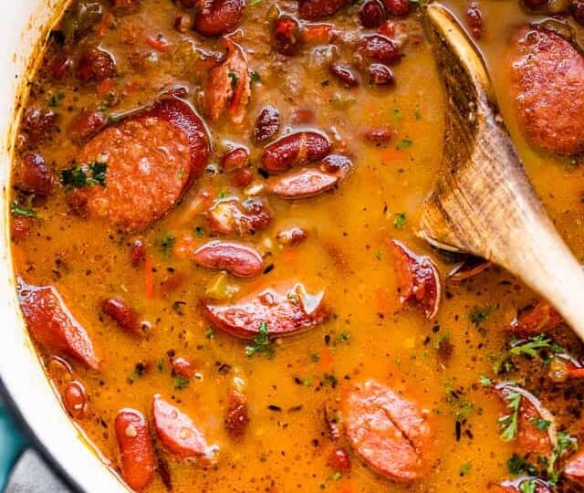 A pot of red beans and sliced sausage in a rich, seasoned broth with a wooden spoon on the side.