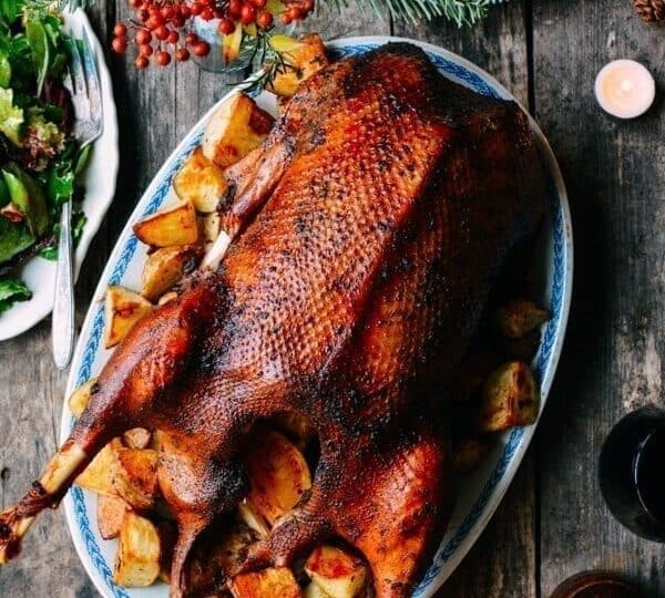 Roast duck garnished with potatoes and herbs on a blue and white platter, surrounded by plates, candles, and a salad on a rustic wooden table.