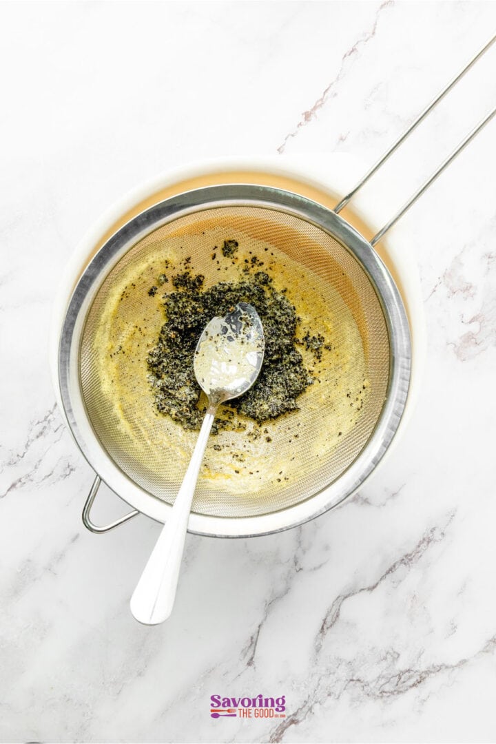 A fine mesh strainer with matcha powder residue sits over a bowl on a marble countertop, alongside a spoon resting inside the strainer. Nearby, a glass of passion fruit juice adds a vibrant splash of color to the serene setting.
