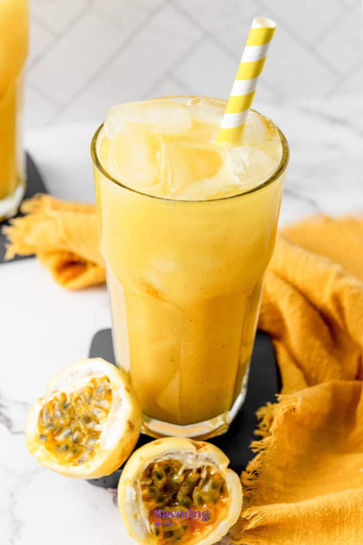 A tall glass filled with a yellow passion fruit juice smoothie, topped with ice cubes and a yellow-striped straw. A halved passion fruit and a yellow cloth are beside the glass.