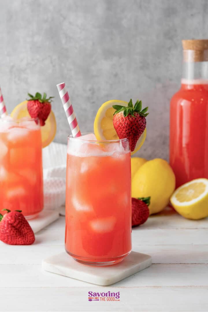 Two glasses of strawberry lemonade with ice, garnished with strawberries and lemon slices, sit on a white surface. A bottle of strawberry lemonade and whole lemons are in the background.