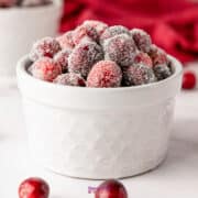 A pristine white bowl brimming with glistening sugared cranberries sits on a white surface, while a vibrant red cloth adds a splash of color in the background.