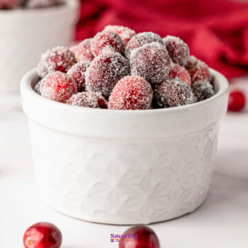 A pristine white bowl brimming with glistening sugared cranberries sits on a white surface, while a vibrant red cloth adds a splash of color in the background.
