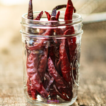A glass jar sits on a wooden surface, brimming with the vibrant hues of drying red chili peppers.