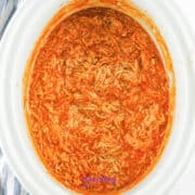 shredded cooked chicken breasts, franks hot sauce, butter and ranch dressing packet cooked to make buffalo chicken dip in a crock pot. The crockpot is white and oval.