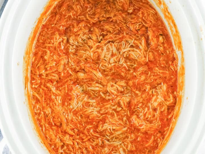 shredded cooked chicken breasts, franks hot sauce, butter and ranch dressing packet cooked to make buffalo chicken dip in a crock pot. The crockpot is white and oval.