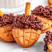 Acorn cookies with chocolate sprinkles and pretzel sticks rest on a marble surface, accompanied by a glass of milk in the background.