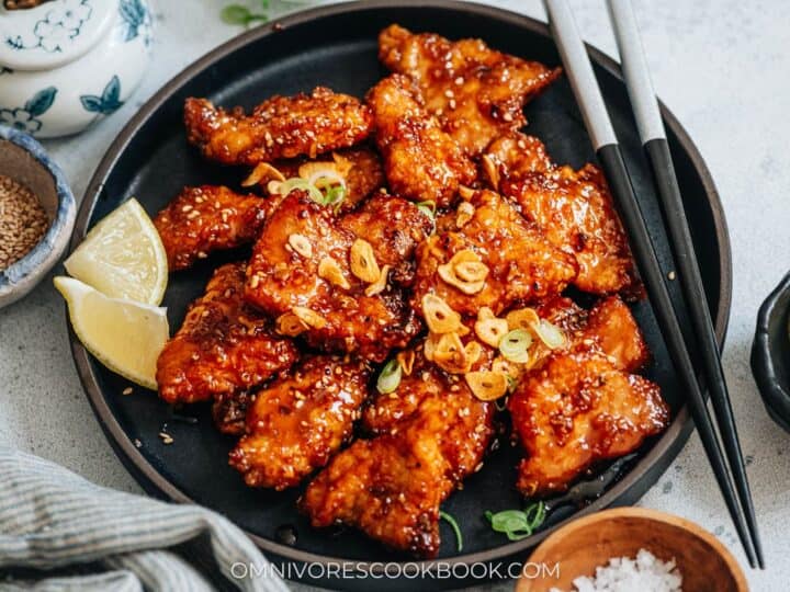 A plate of crispy fried chicken pieces glazed with sauce, garnished with sliced garlic and green onions. Lemon wedges are placed on the side, with chopsticks resting on the plate.