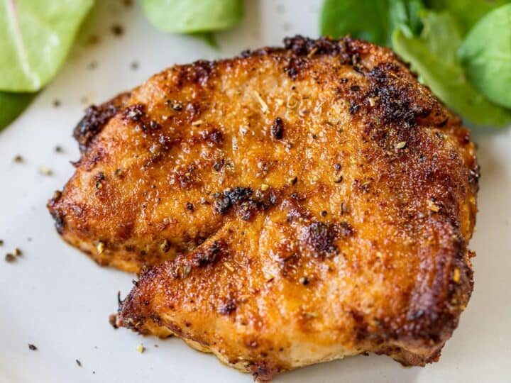 A piece of seasoned, grilled chicken breast on a white plate with fresh green salad leaves in the background.