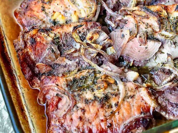 Close-up of roasted lamb chops topped with sliced onions and herbs in a glass baking dish.