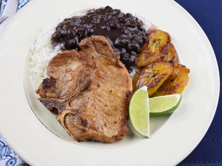 A plate with grilled pork chop, white rice topped with black beans, slices of fried plantains, and lime wedges.