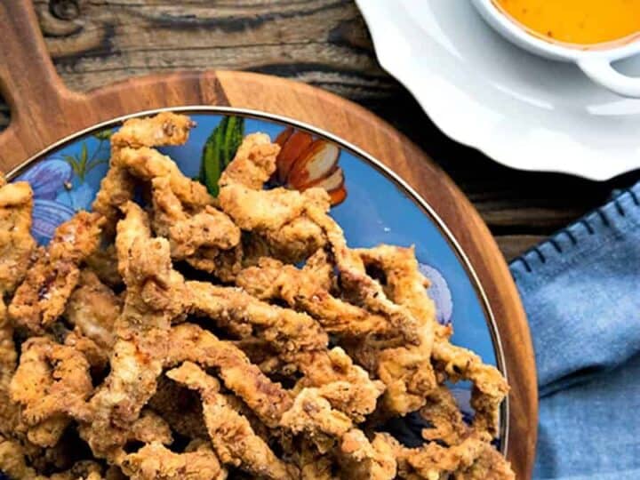 Fried chicken strips on a decorative plate with a bowl of reddish-orange dipping sauce on a wooden surface.