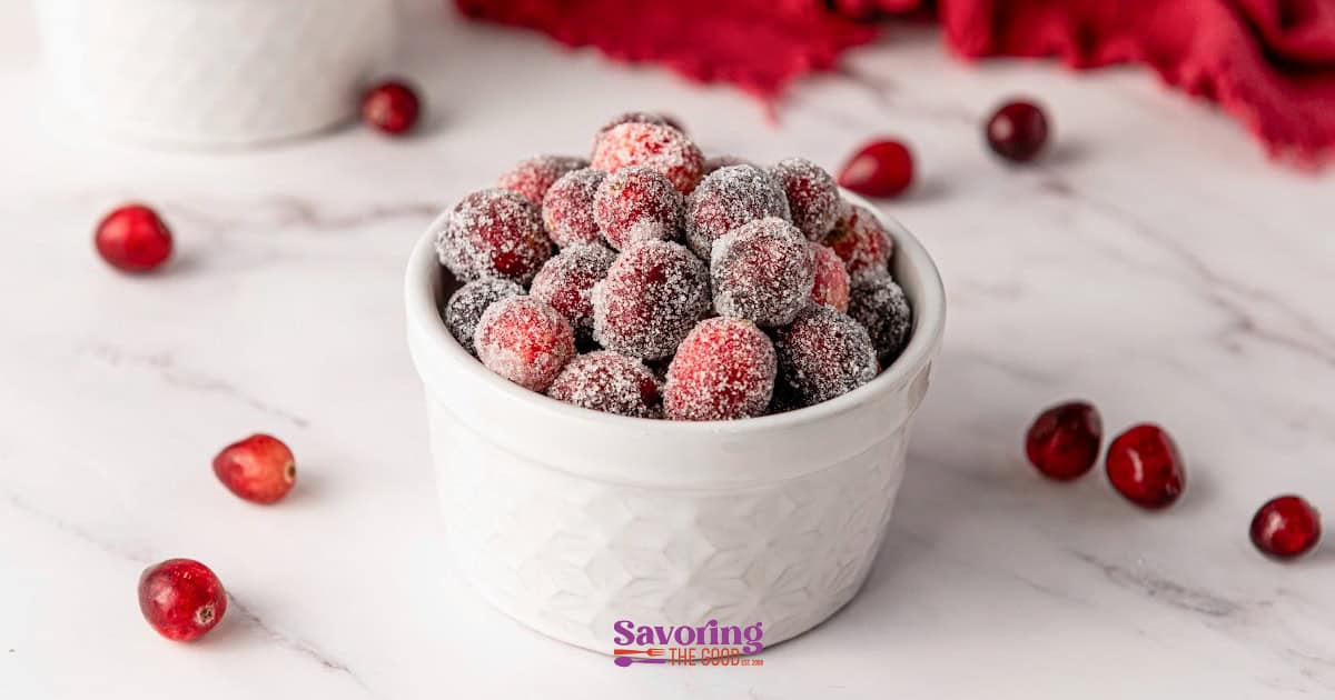 A white bowl brimming with glistening sugared cranberries sits elegantly on a marble surface, with a handful of the vibrant berries playfully scattered around.