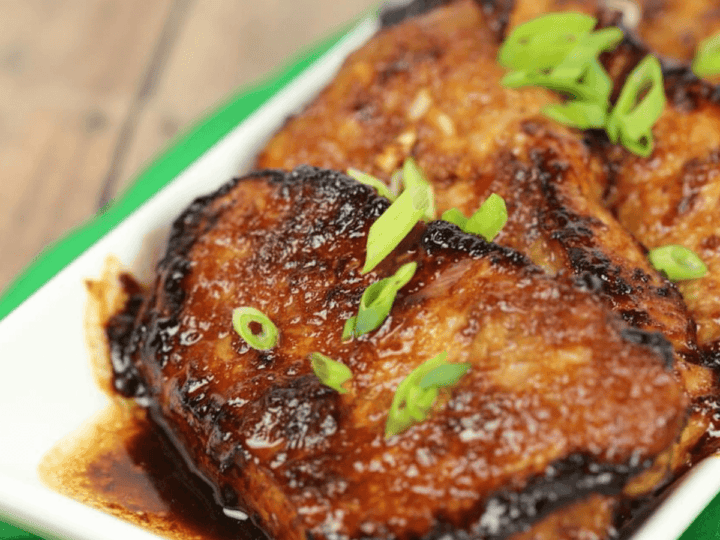 Grilled chicken pieces glazed with sauce, garnished with sliced green onions, served in a white dish on a green cloth.