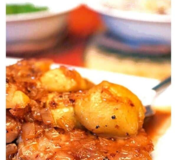 Close-up of pan-seared pork chops topped with caramelized onions and potatoes, with a backdrop of blurred dishes. Text reads "Harvest Pork Pan-Seared Pork Chops.