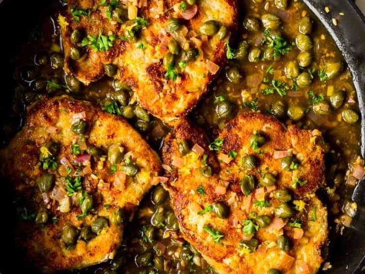 A skillet with three breaded pork chops topped with capers and herbs, surrounded by a sauce, placed on a wooden board with a striped cloth.