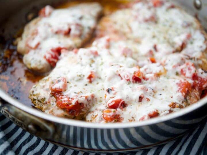 A skillet with chicken breasts topped with melted cheese and diced tomatoes.
