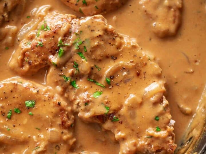 Pork chops in a creamy brown gravy with chopped herbs on top, in a skillet.