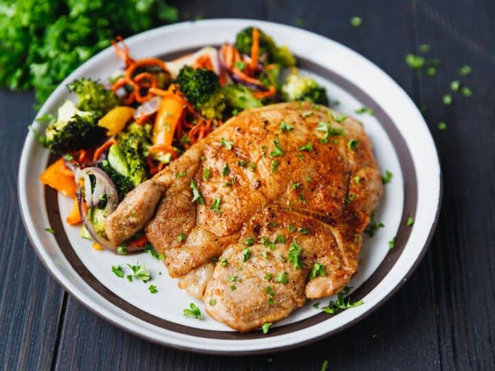 Grilled chicken breast garnished with parsley, served with mixed vegetables including broccoli and bell peppers on a white plate.