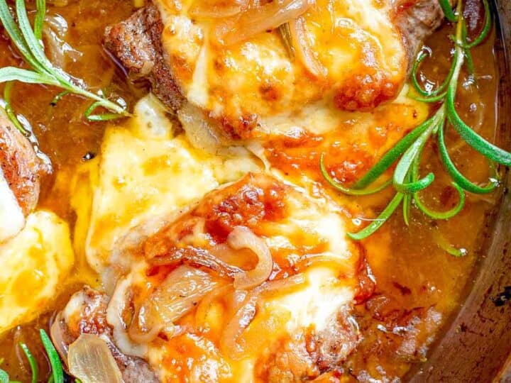 Close-up of a skillet with cooked meat topped with melted cheese and caramelized onions, garnished with sprigs of fresh rosemary.