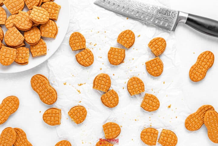 Halved peanut-shaped cookies scattered on parchment paper, resembling charming acorn cookies, with a knife nearby and a plate of whole cookies.