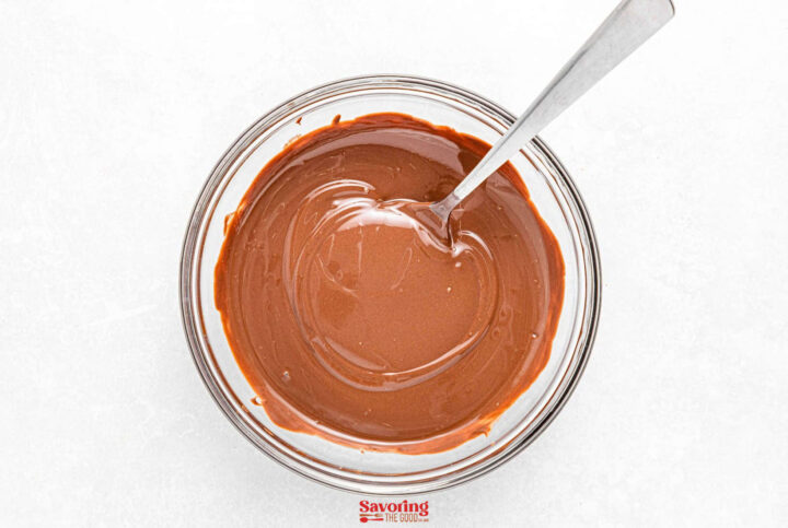 A glass bowl brimming with smooth, melted chocolate, perfect for drizzling over your favorite treats, with a spoon resting inside. Ideal for elevating your acorn cookies recipe. Set against a pristine white background, it invites indulgence and creativity in equal measure.